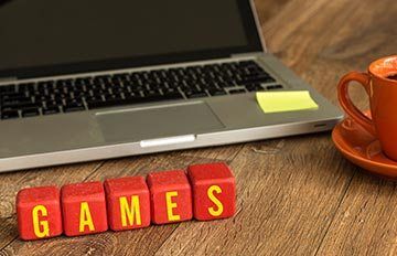 five red cubes with the word "games" in white on them. Behind them is a laptop and next to them is a mug of coffee