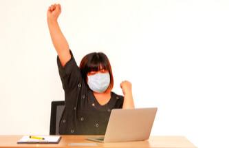 a very happy, winning woman wearing a surgical mask playing on her laptop 