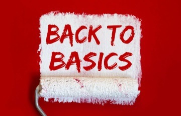 A paint roller painting white paint on a red wall with the stenciled words ‘back to basics’ in red