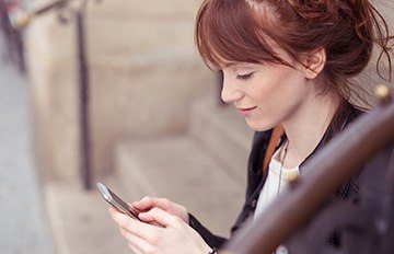 a pretty red haired young woman sitting on her front steps using her mobile device for communication or gaming