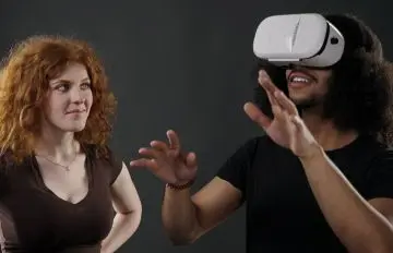 a redhead young woman looks on as her boyfriend or husband experiences Virtual Reality with a headset
