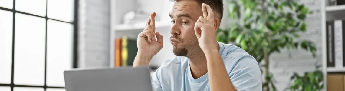 a young man with a short beard and moustache crossing his fingers while looking at his laptop maybe playing online casino games