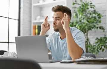 a young man with a short beard and moustache crossing his fingers while looking at his laptop maybe playing online casino games