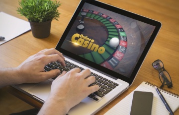 man's hands at laptop playing roulette at online casino. On the table are a small potted plant, pad and pen, glasses, and phone