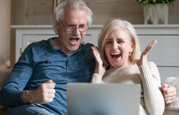 very happy white haired older couple playing at an online casino while comfortably sitting on the sofa.