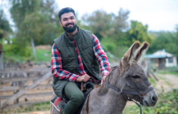 30-something man riding a donkey for fun at a donkey ranch. He has a short beard and moustache and is wearing a flannel shirt