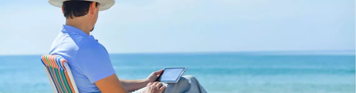 a barefoot man wearing a hat sitting on a folding chair at the ocean palying on his mobile device