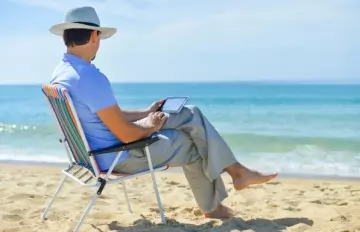 a barefoot man wearing a hat sitting on a folding chair at the ocean palying on his mobile device