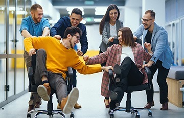 six office workers having fun in the hall with two sitting in wheeled chairs and the others pushing them through the hall