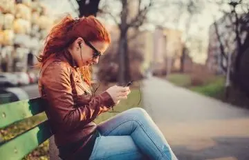 a young woman with curly red hair sitting on a park bench in a park like Central Park accessing her mobile device