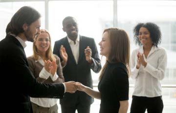 new employee being hired by an officer of the company with a handshake and cheered on by her colleagues and peers
