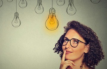 attractive young woman with short, curly brown hair looking up at light bulbs with one lit up to signify creativity