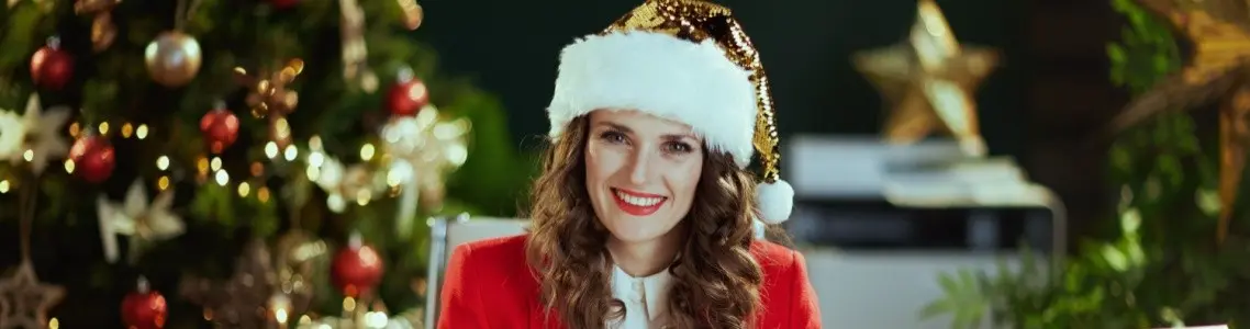 a smiling woman at her desk with her laptop open, with a Santa hat on, and a well lit Christmas tree in the background