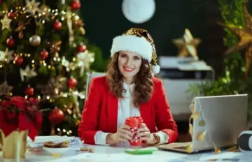 a smiling woman at her desk with her laptop open, with a Santa hat on, and a well lit Christmas tree in the background