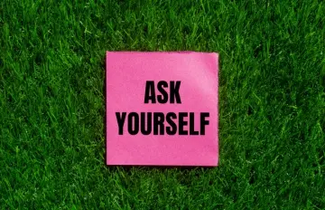 a sign on pink paper set against a green, synthetic grass background with black letters saying "Ask Yourself"