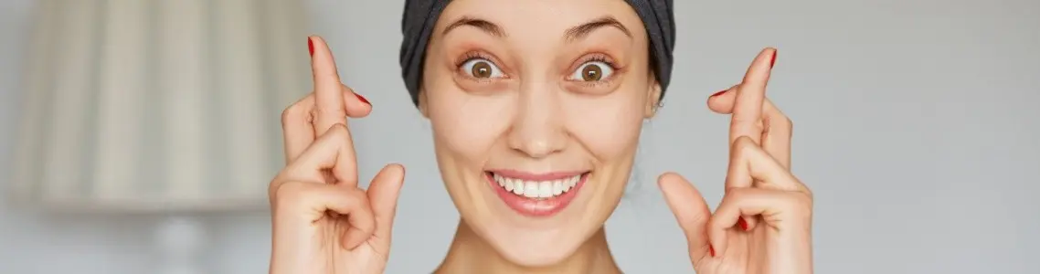 a young woman with her fingers crossed making an exaggerated smile at the camera as if she is making fun of superstition