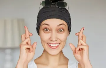 a young woman with her fingers crossed making an exaggerated smile at the camera as if she is making fun of superstition