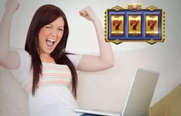 a young woman excitedly cheering her win playing an online slot.  She is sitting on the sofa with her laptop on her lap.
