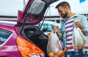 a man loading groceries into the trunk of his small, purple car.  The car and easy shopping are examples of modern convenience.