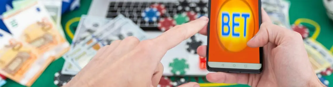 man at a table at home holding a smartphone, the word bet on the screen of his laptop, and casino chips all around the laptop