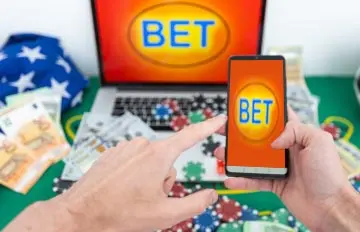 man at a table at home holding a smartphone, the word bet on the screen of his laptop, and casino chips all around the laptop